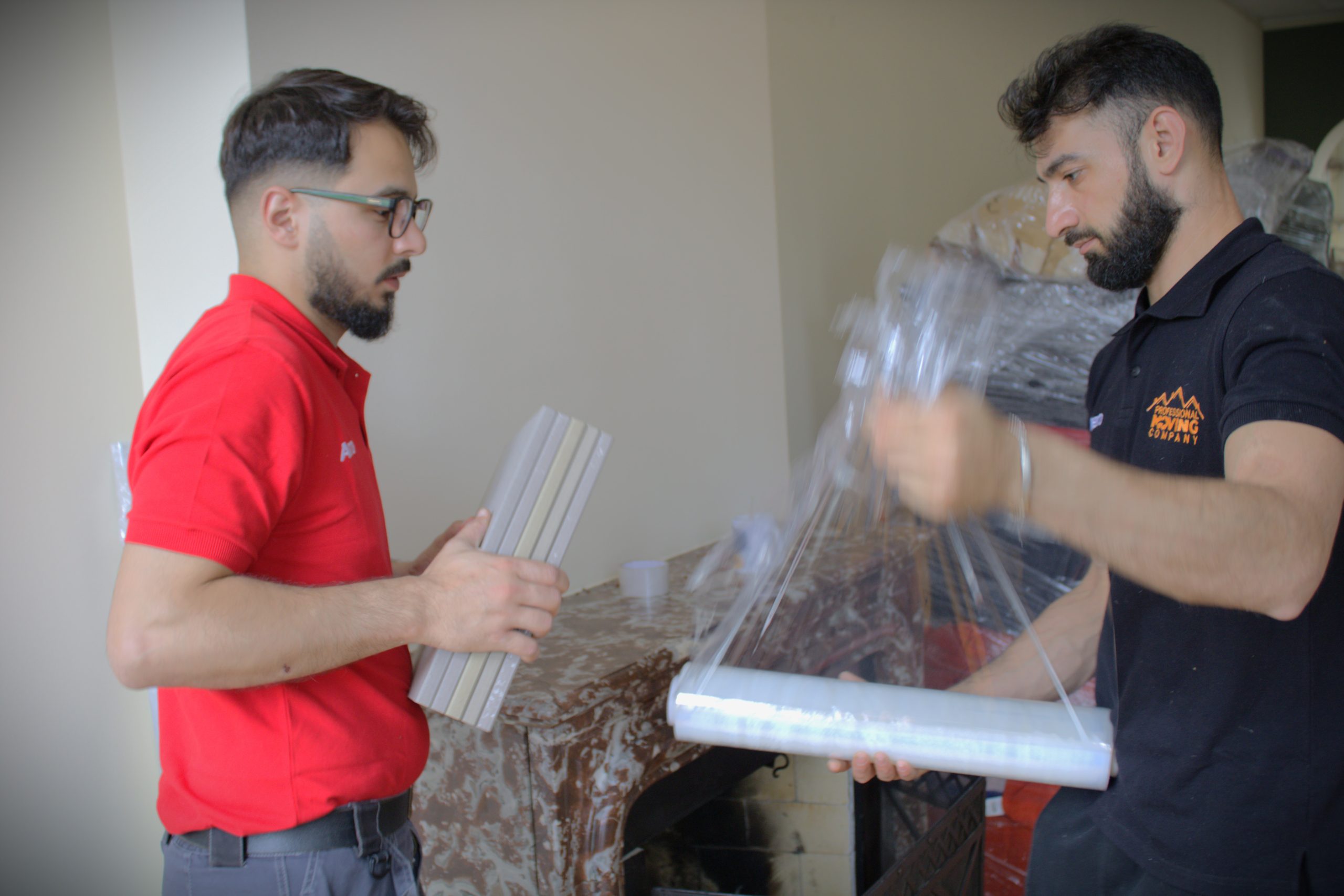 Two movers from Professional Moving Company preparing items for a move. One mover is holding packaging materials, while the other is wrapping items with plastic wrap.