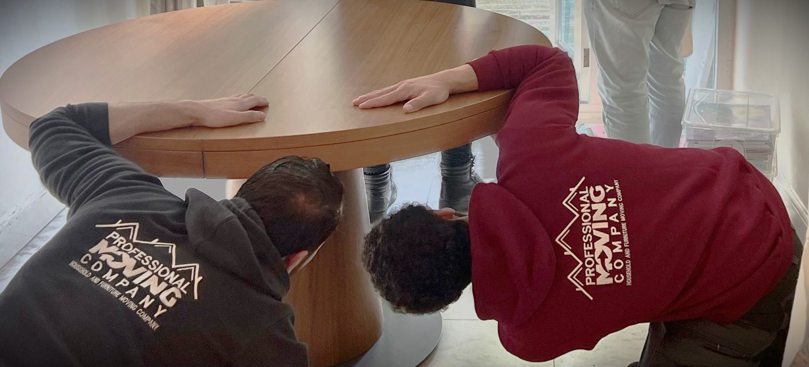 Two movers from Professional Moving Company adjusting a wooden table. They are crouched under the table, ensuring it is securely assembled.