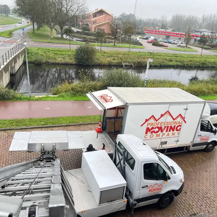 Twee verhuiswagens van "Professional Moving Company" staan geparkeerd naast een kanaal, met een verhuislift in gebruik op een geplaveide straat.